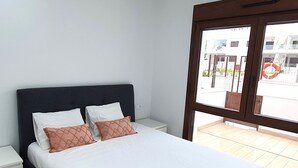 Une chambre avec un grand lit, du linge de lit blanc, une tête de lit sombre, une table de chevet et une vue sur l’extérieur, accessible par une porte vitrée.