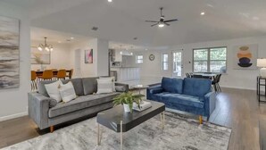 Amplia sala de estar con un sofá gris, un sofá azul y una mesa de centro de mármol. Se observa un comedor con mesa y sillas, y una cocina al fondo.