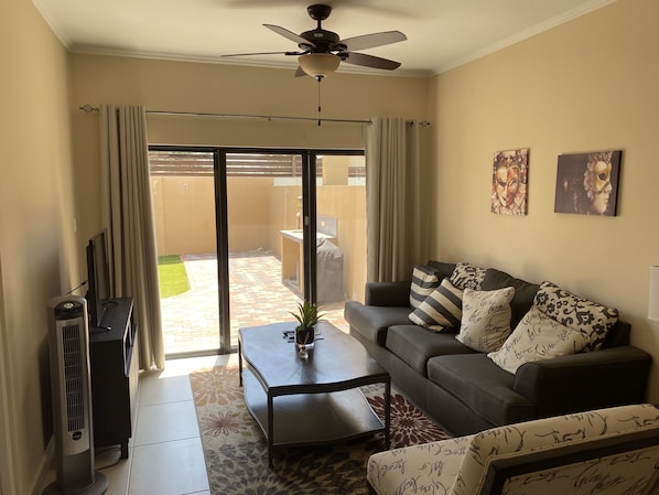 A living room with a sofa, coffee table, and a ceiling fan. There is a sliding glass door leading to a patio with a tiled floor and a small garden.