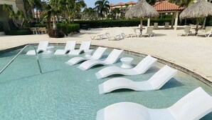 A swimming pool with clear water, surrounded by white lounge chairs and a thatched umbrella structure.