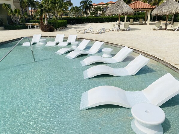 A swimming pool with clear water, surrounded by white lounge chairs and a thatched umbrella structure.