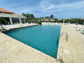 A swimming pool with lounge chairs and umbrellas, surrounded by palm trees and buildings.