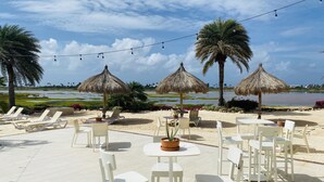 An outdoor seating area with white chairs and tables, thatched umbrellas, and palm trees.