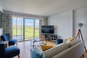 Una sala de estar moderna con un televisor grande, asientos cómodos y vistas a un paisaje verde a través de puertas de vidrio.