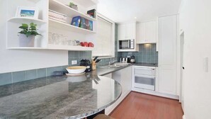 A compact kitchen with white cabinets, a microwave, and a sink. There are shelves with books and a potted plant.