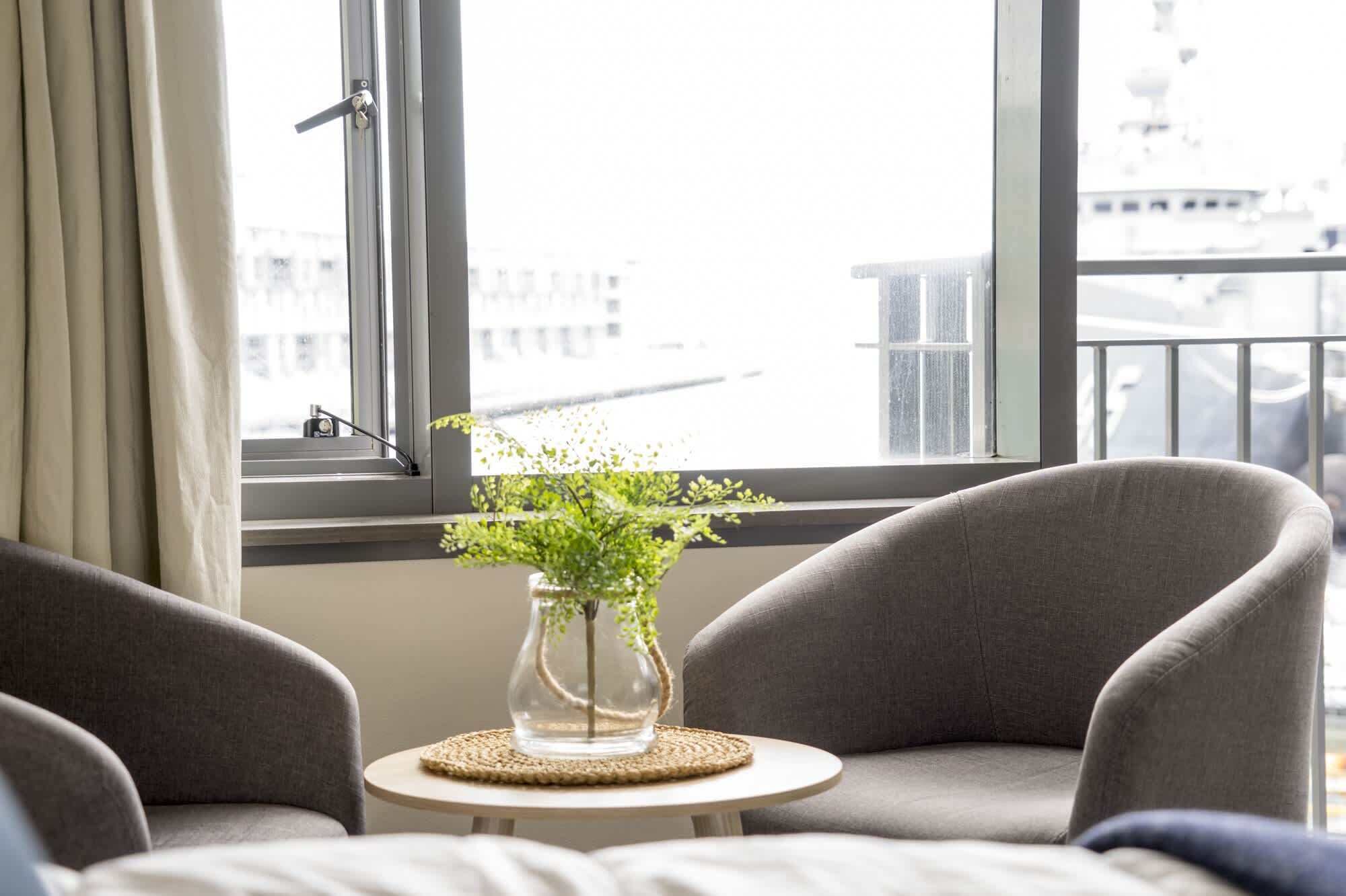A room with two grey armchairs, a small round table with a vase of green plants, and a view of buildings through large windows.