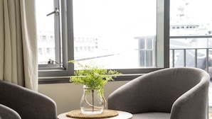 A room with two grey armchairs, a small round table with a vase of green plants, and a view of buildings through large windows.