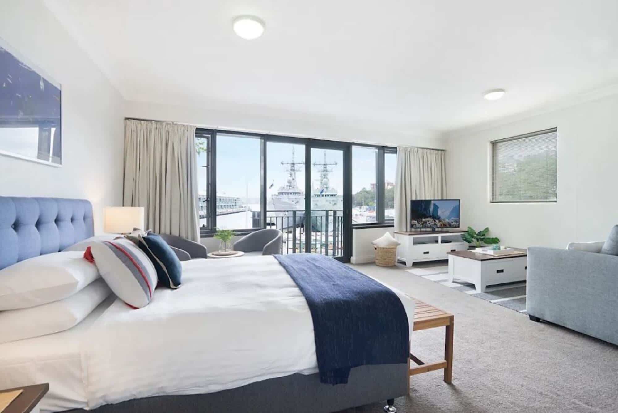 A modern hotel room with a large bed, a TV, a balcony with a view of the sea, and a seating area with a table and chairs.
