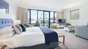 A modern hotel room with a large bed, a TV, a balcony with a view of the sea, and a seating area with a table and chairs.