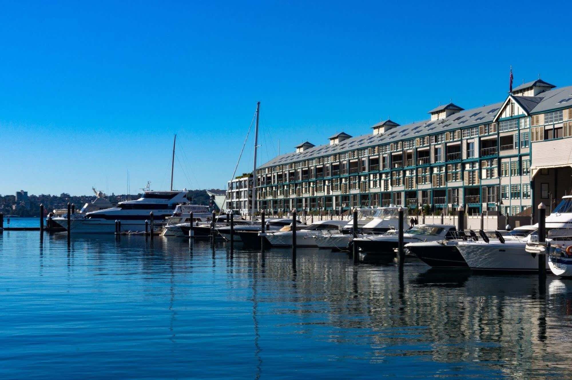 A marina with multiple yachts docked, a large building with multiple balconies, and a clear blue sky.
