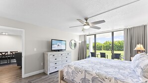 A bedroom with a bed, a ceiling fan, a television, a dresser, and a view of a garden through large windows. - Cape Canaveral Charm Double Wide Patio & Heated Amenities (Cape Canaveral)