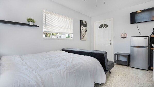 A bedroom with a bed, a small desk, a refrigerator, and a TV mounted on the wall.