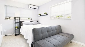 Camera da letto con un letto, un divano grigio, una finestra con tende, uno scaffale con piante e un condizionatore d'aria.