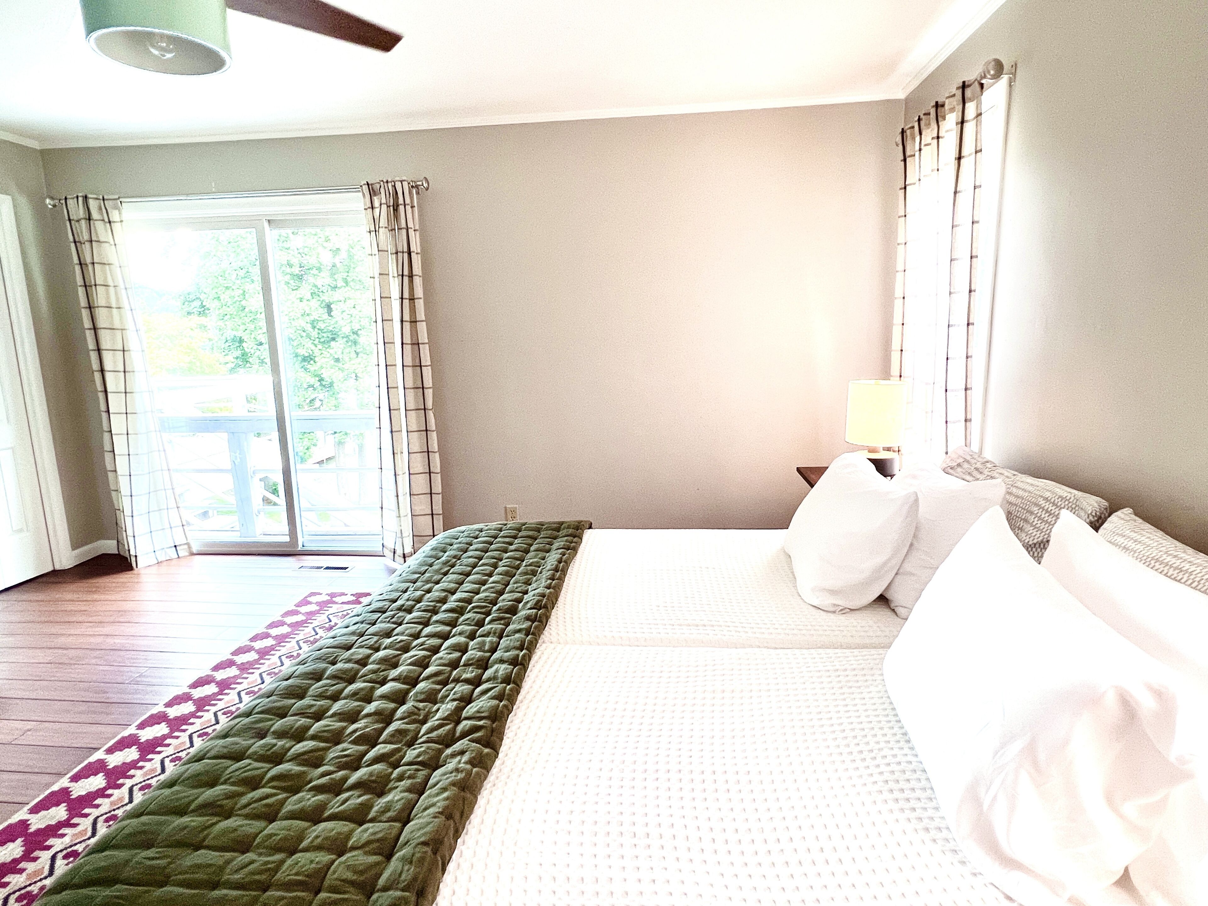 A hotel room with a bed, a nightstand, a lamp, and a view of the outdoors through a window with curtains.