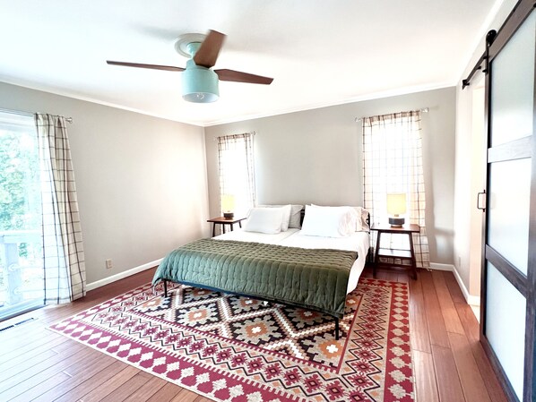 Une chambre à coucher comprenant un lit, un ventilateur de plafond, deux tables de chevet avec des lampes, un tapis à motifs et une grande fenêtre avec des rideaux.
