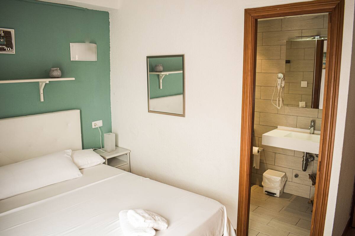 Une chambre d’hôtel avec un grand lit, une table de chevet, un miroir, une étagère avec des objets décoratifs et une vue sur la salle de bain, où l’on aperçoit un lavabo et une douche.
