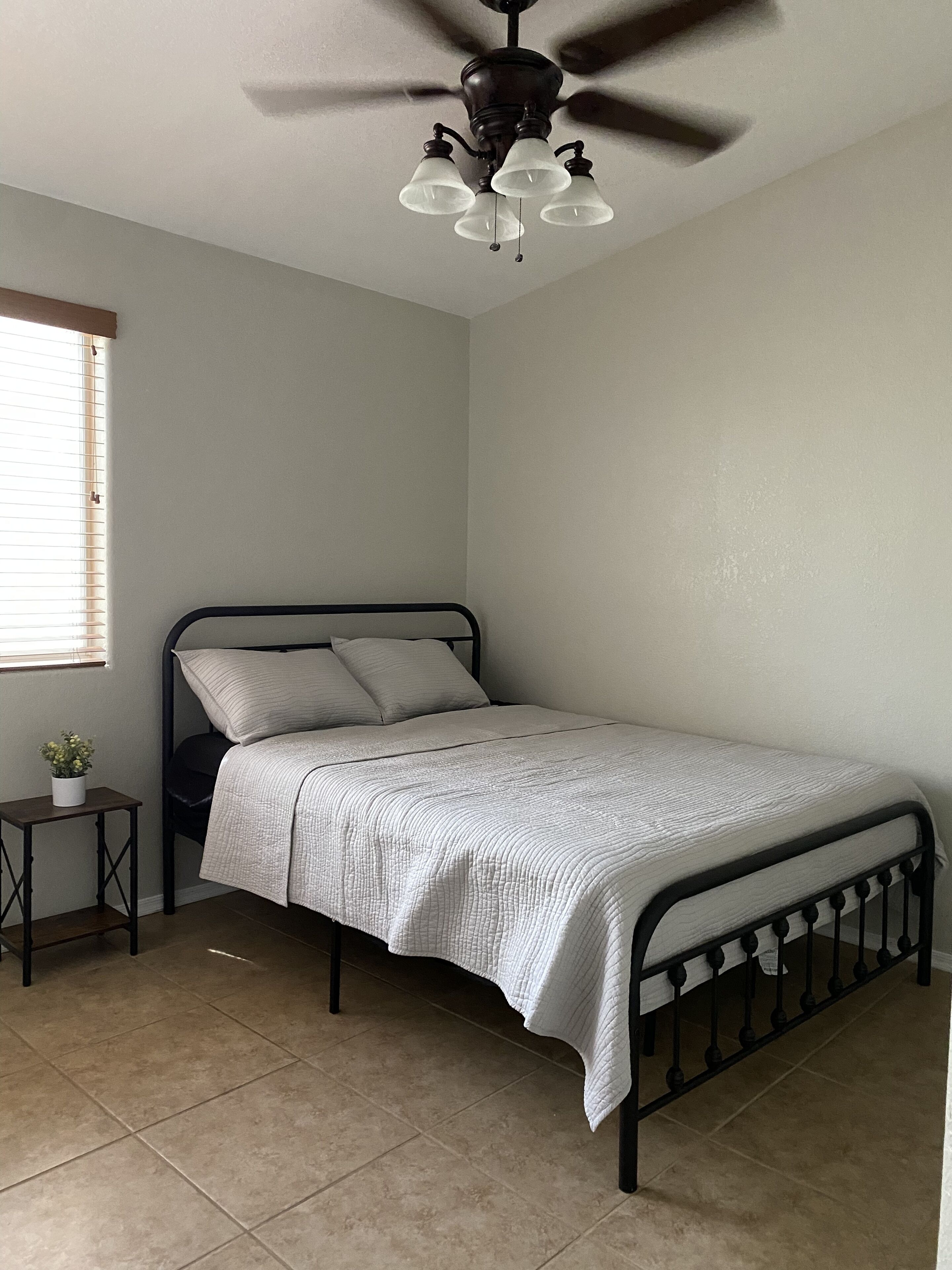 A bedroom with a single bed, a small bedside table, a ceiling fan, and a window with blinds.