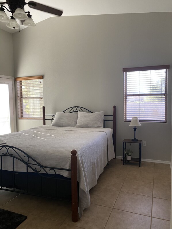 A bedroom with a bed, a ceiling fan, a window with blinds, and a nightstand with a lamp.