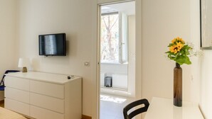 A hotel room with a flat-screen TV mounted on the wall, a white dresser, a small table with a vase of sunflowers, and a chair. - Pisacane Suite (Salerno)