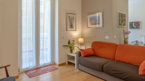 A living room with a grey and orange sofa, a small white side table with a lamp, and a vase with flowers. - Pisacane Suite (Salerno)
