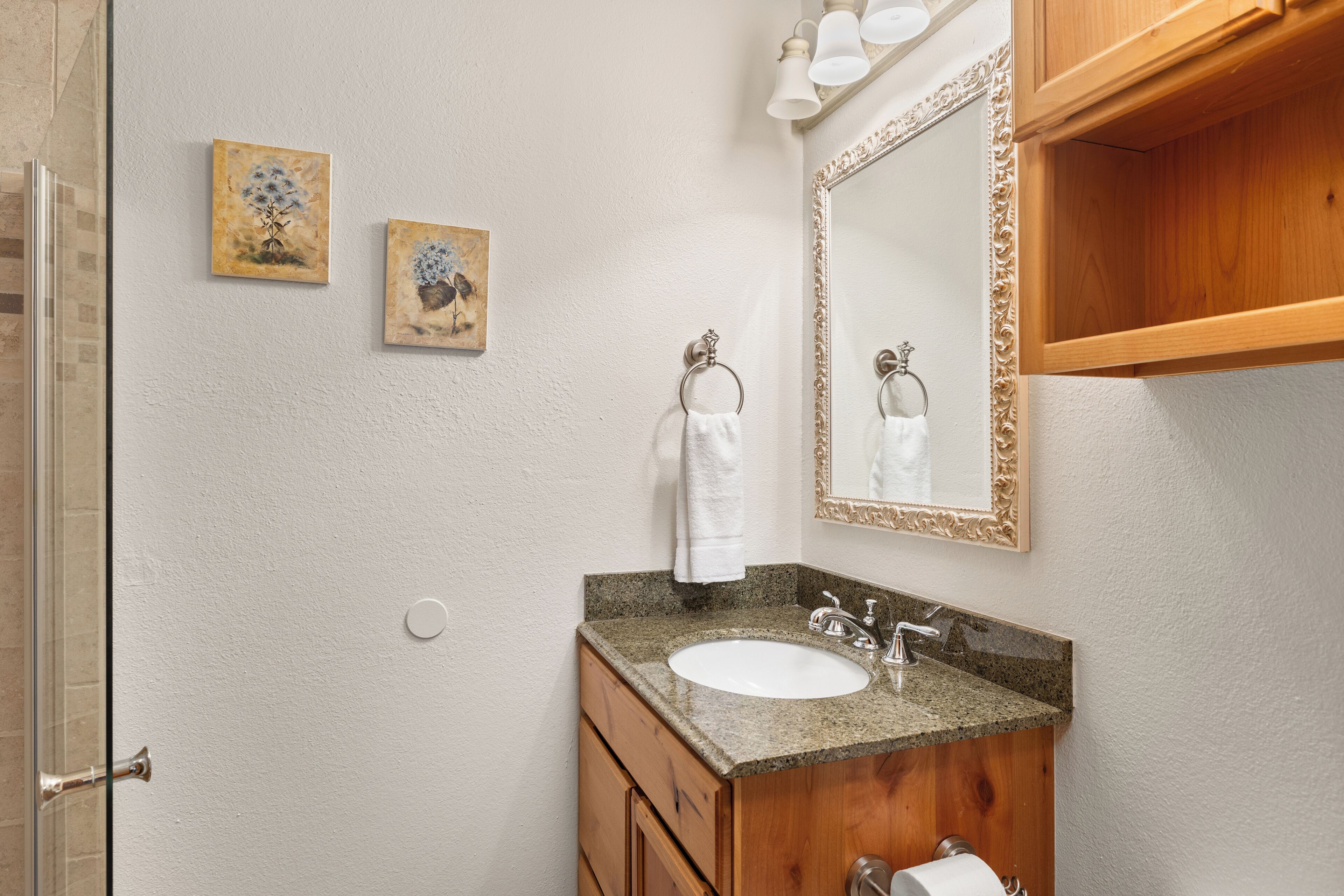 A bathroom with a granite countertop sink, wooden vanity, two framed floral paintings, a mirror with a decorative frame, and a towel rack.