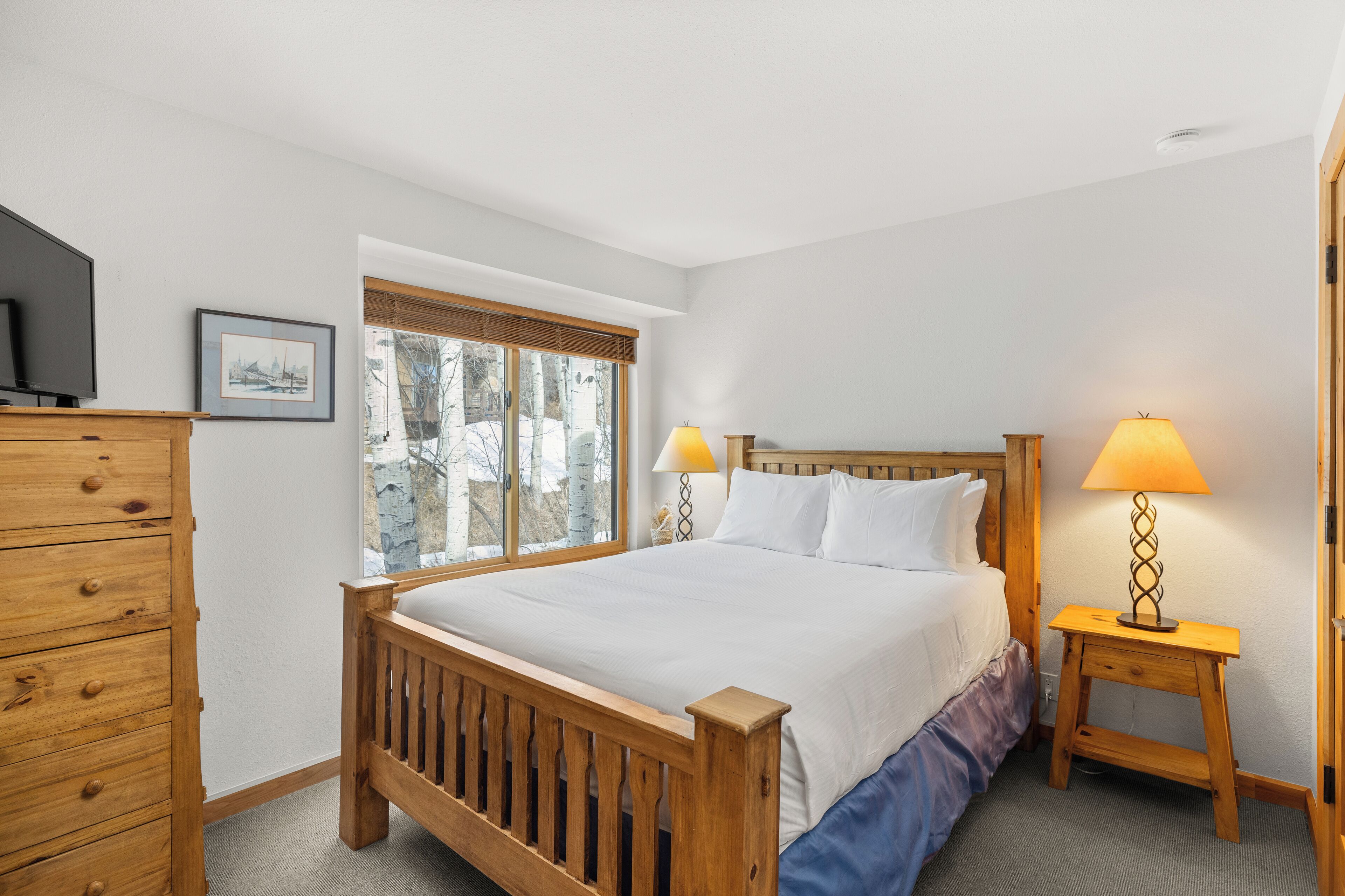 Un dormitorio con una cama de madera, dos lámparas de noche, un aparador y una ventana con vista a árboles.