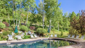 A swimming pool with lounge chairs and umbrellas, surrounded by trees and a hilly landscape. - Seasons Four by Snowmass Vacations (Snowmass Village)