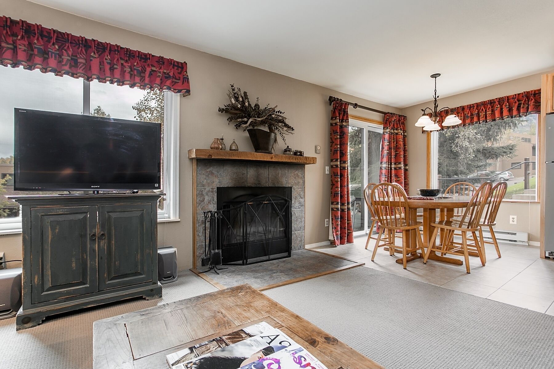 A living room with a fireplace, a dining area with wooden chairs, a television, and a large window with curtains.