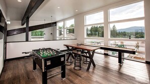 A spacious room with a foosball table, a long wooden table, and multiple bar stools. Large windows provide a view of the outdoors. - Beautiful 2BD + 2BA Home! King Beds! Hot Tub! Pool! Walk to downtown. (Whitefish)