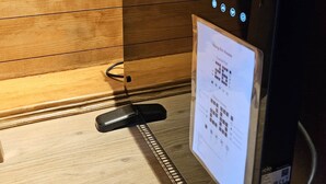 Digitaalinen kello, jonka taustana on puupaneeli, ja sen yhteydessä on ohjauspaneeli sekä lattialla kaukosäädin.