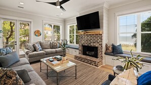 A modern living room with a fireplace, large windows, and a ceiling fan. - Majestic Oaks, Breathtaking Sunrise Ocean Views, Shared Pool, Near the Village (St Simons Island)