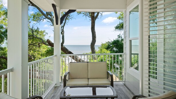 A covered porch with wicker furniture, a glass table, and a view of the ocean. - Majestic Oaks, Breathtaking Sunrise Ocean Views, Shared Pool, Near the Village (St Simons Island)