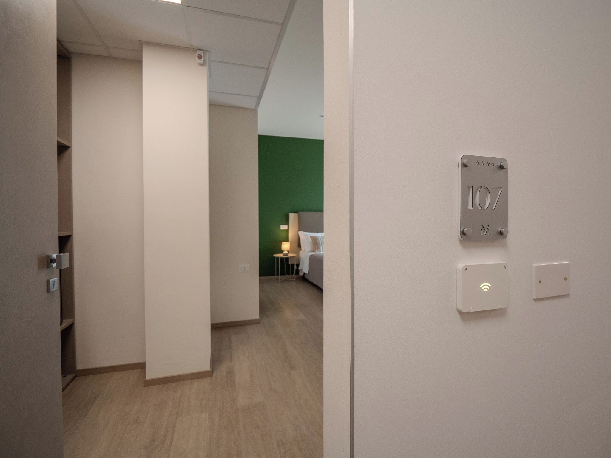 Habitación de hotel con una pared de acento verde, una cama, una mesita de noche y un panel de control en la pared.