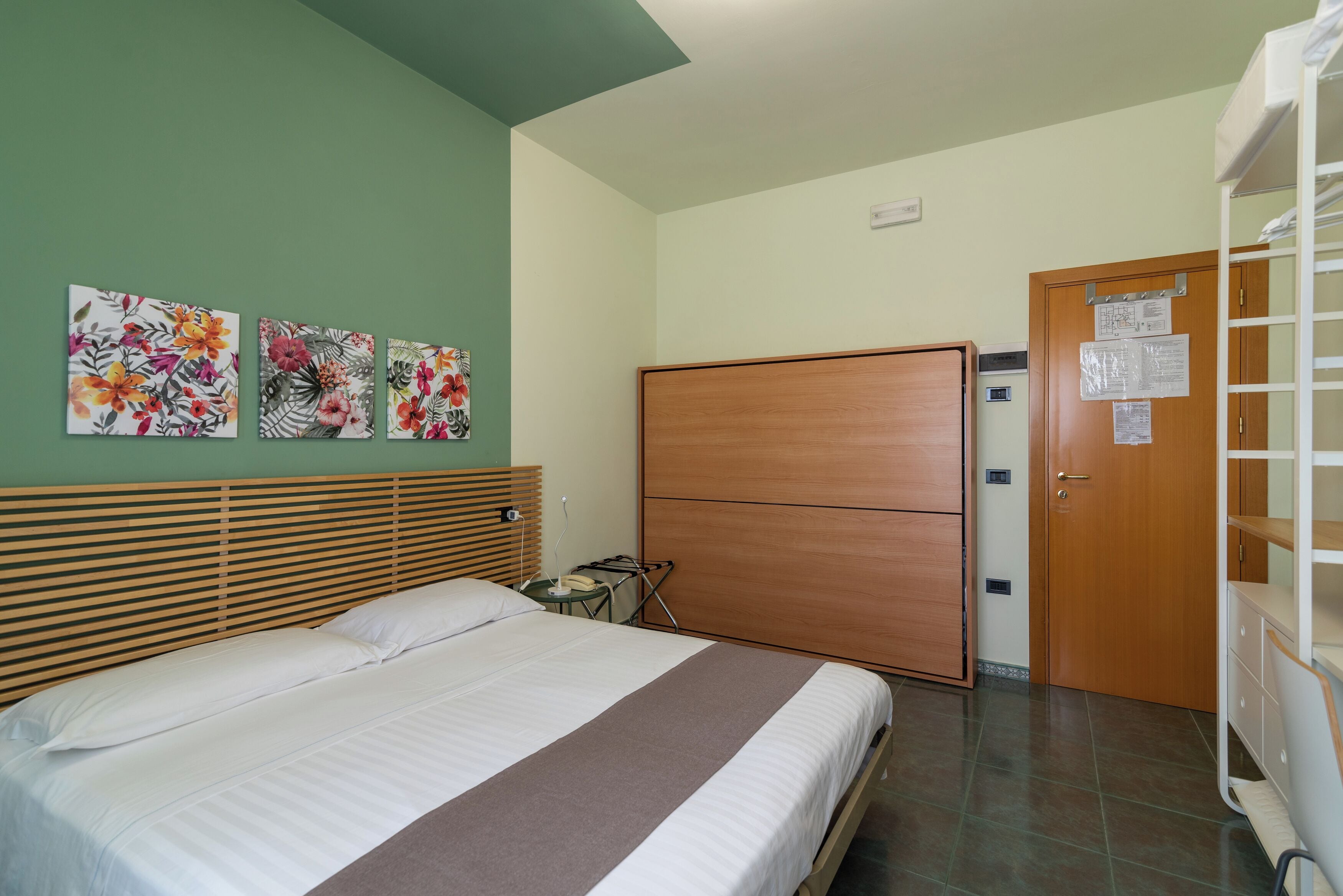 A hotel room with a bed, a wooden headboard, a green wall, and three floral paintings.