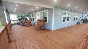 A covered outdoor deck with wooden flooring, a seating area with chairs, and a view of a building with windows. - Flying Fish Yellow Fin-Beach Access-Island Realty (Grand Isle)