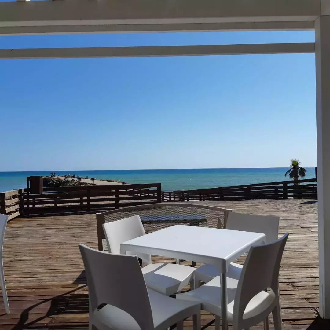 A covered outdoor seating area with white tables and chairs, overlooking a beach and the ocean.