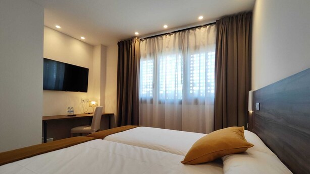 Una habitación de hotel con dos camas, un escritorio, una silla, un televisor y una ventana con cortinas.