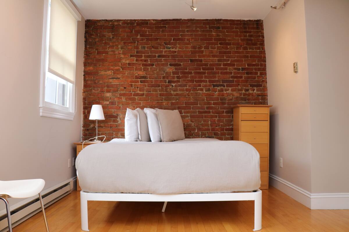 A bedroom with a brick wall, a bed, a nightstand, a dresser, and a window.