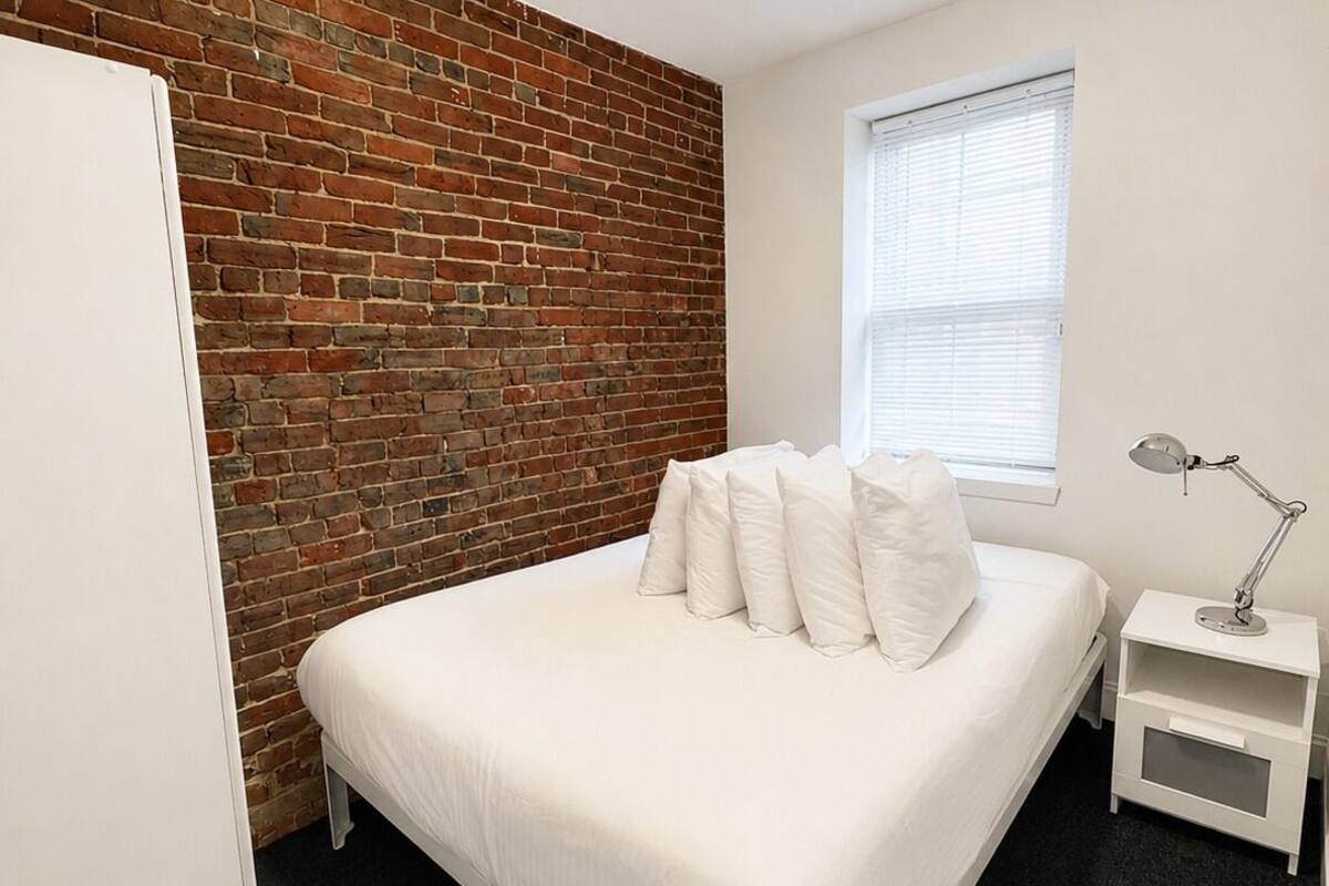 A bedroom with a brick wall, a white bed, a window with blinds, a bedside lamp, and a ceiling light.
