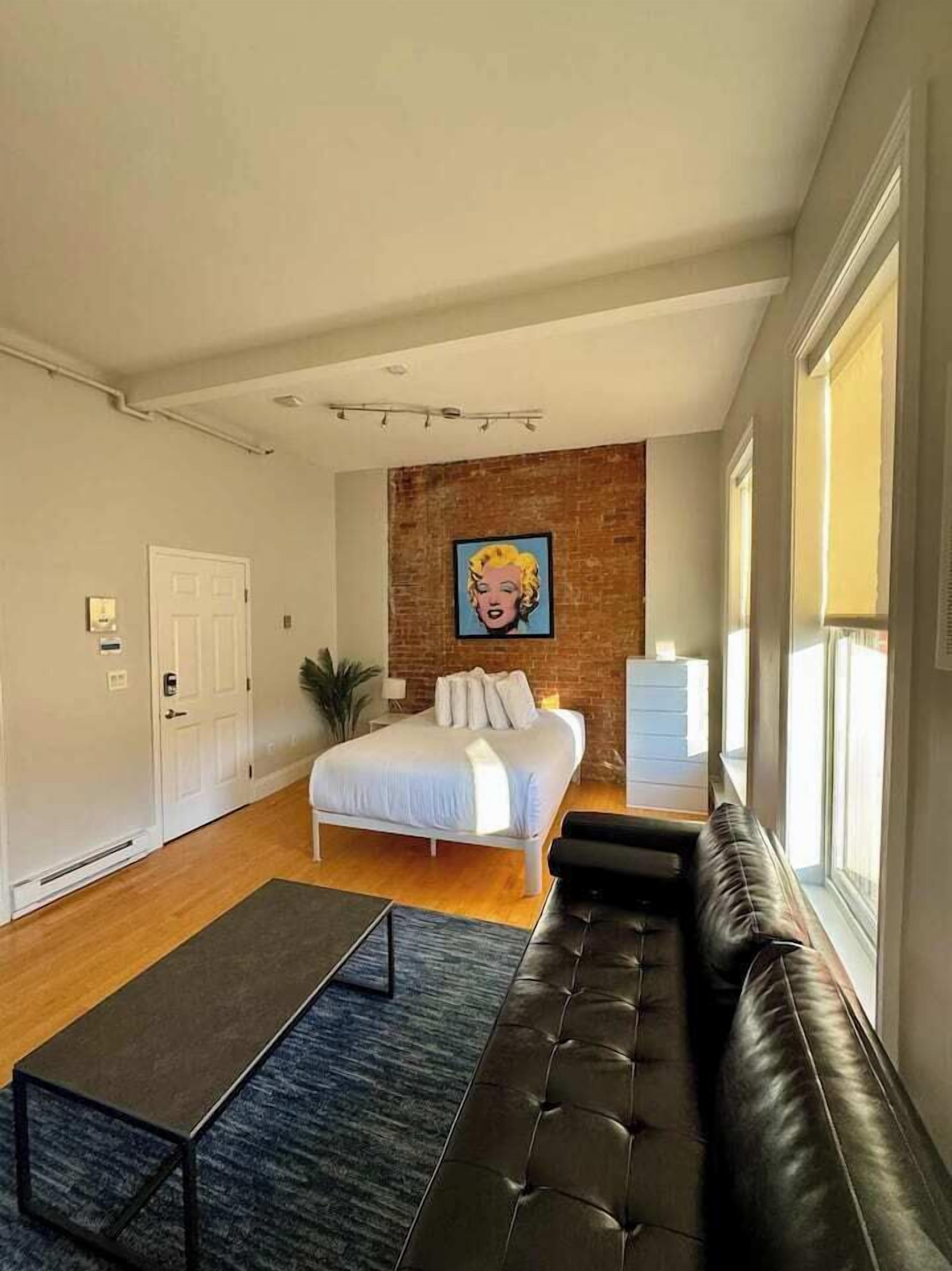 Un dormitorio con una cama, un sofá de cuero negro, una mesita, una pared decorativa de ladrillo y una ventana.