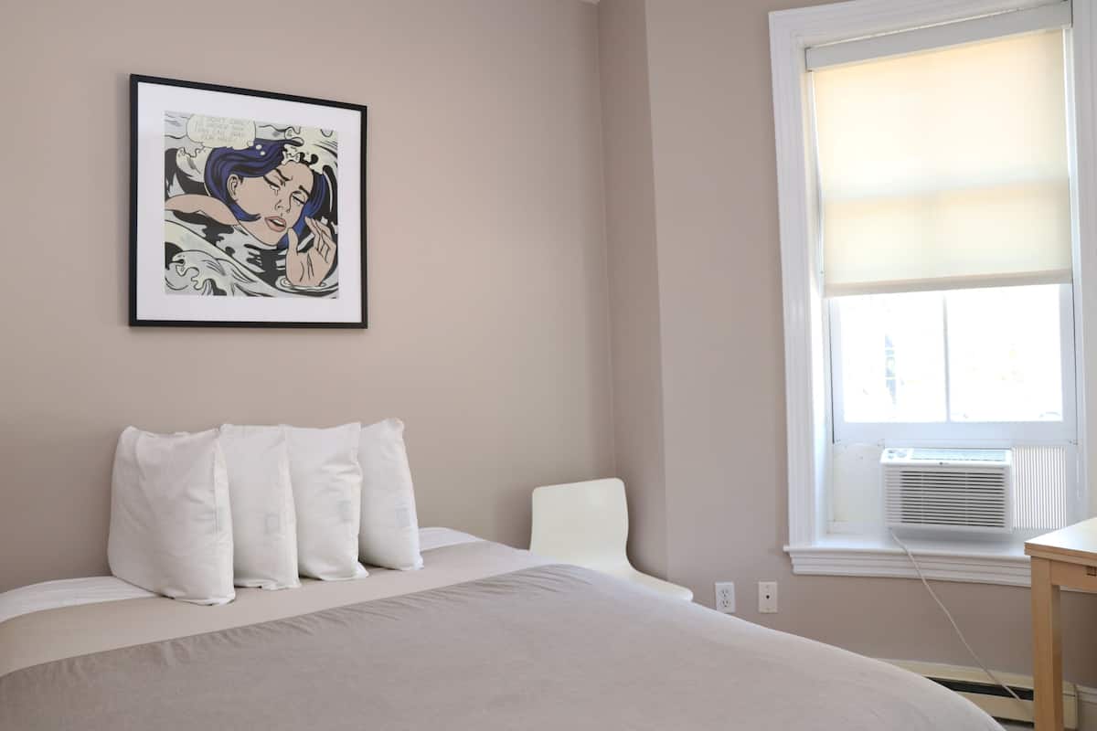 A bedroom with a bed, pillows, a framed picture, a window, and an air conditioning unit.