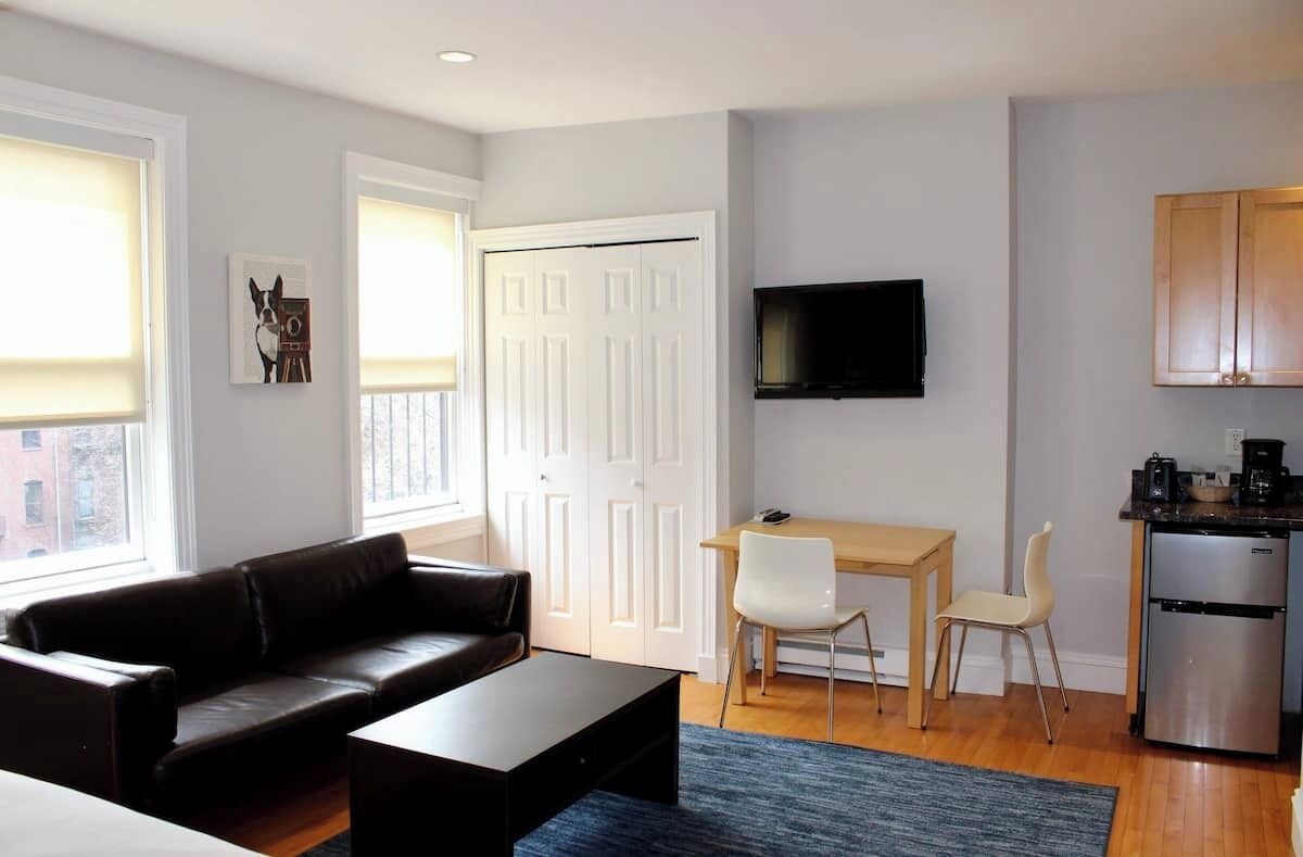 Una sala de estar con sofá, mesa de centro, televisor montado en la pared, mesa de comedor con sillas, y una zona de cocina con armarios y refrigerador.