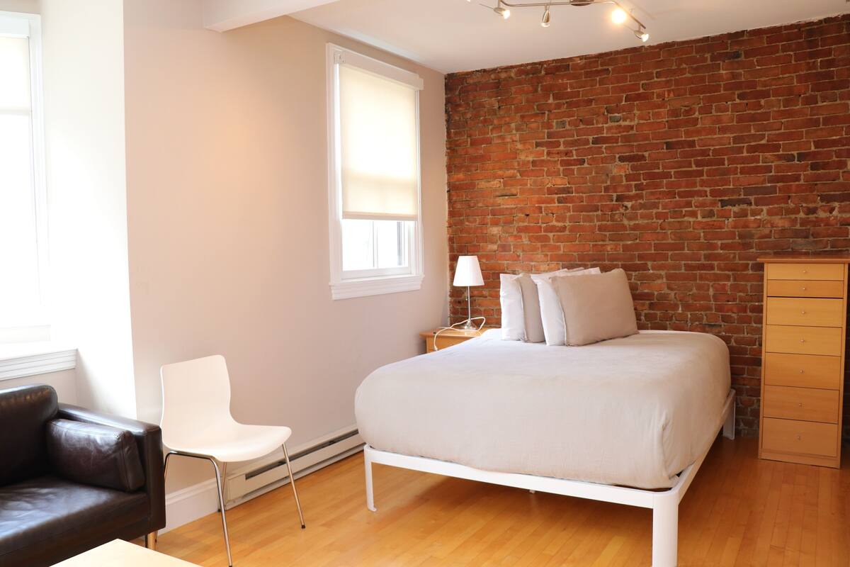 A bedroom with a bed, a chair, a dresser, a lamp, and a brick wall.