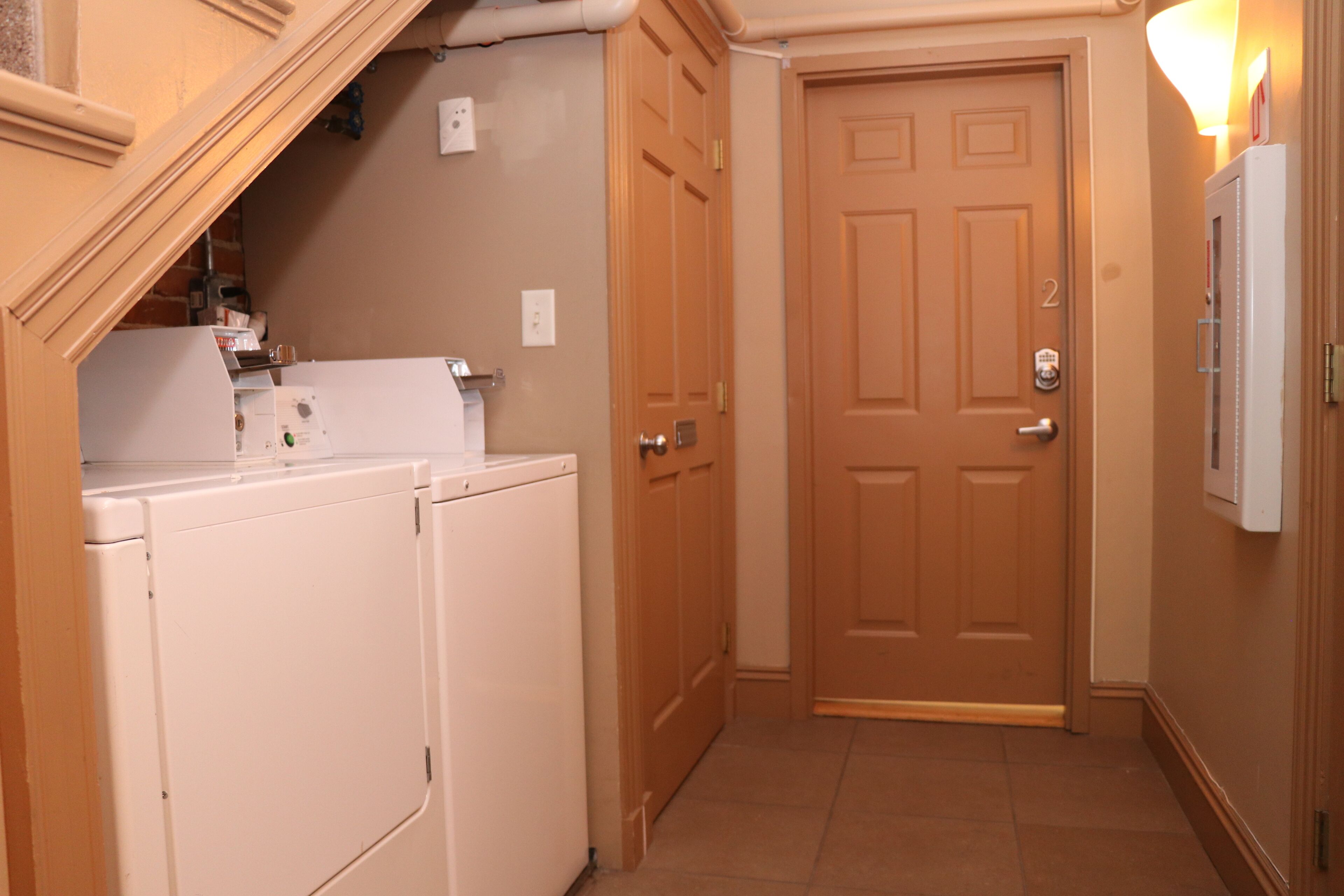 A laundry room with two white washers and dryers, a wooden door, and a light fixture on the wall.