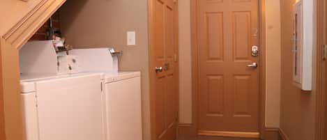 Una sala de lavandería con dos lavadoras y secadoras blancas, una puerta de madera y una lámpara en la pared.