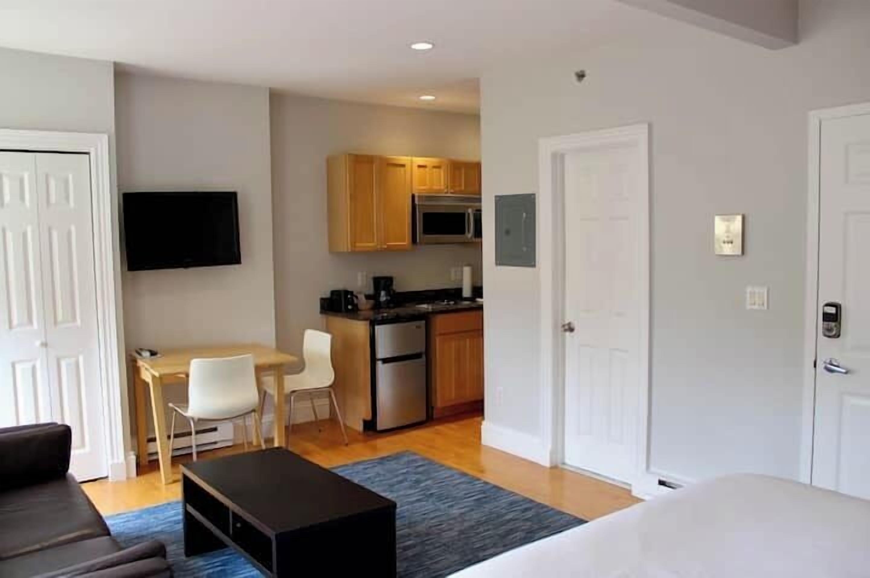 A compact hotel room with a bed, a small desk, a chair, a television, and a kitchenette.