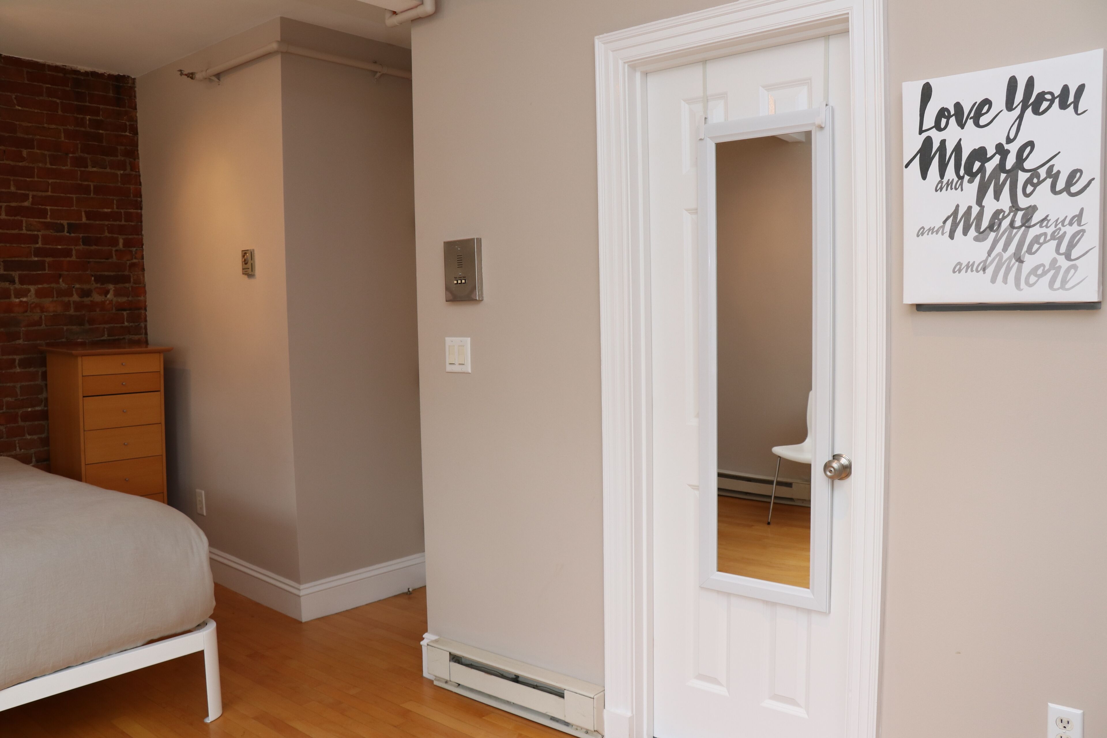 Quarto com cama, penteadeira, porta branca e placa na parede.