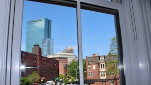 A view of a cityscape through a window, including a tall glass building, brick buildings, and a street with parked cars and a bus stop. - Maverick Suites at 402 Columbus Ave (Boston)