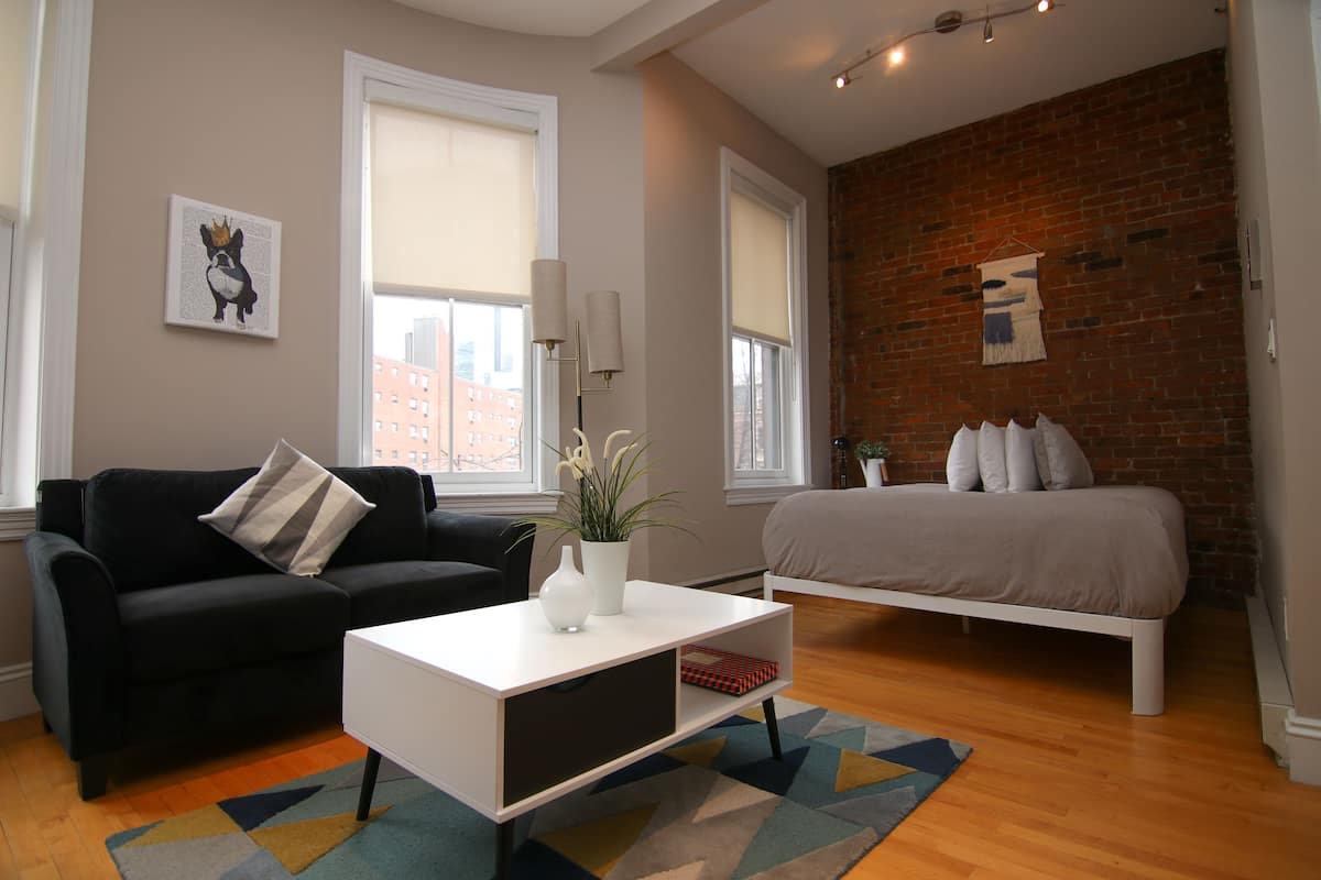 A modern bedroom with a black sofa, a white bedside table, a bed, and a brick wall.