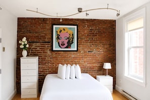 A bedroom with a brick wall, a bed with white bedding, a dresser, a nightstand, and a framed picture. - Maverick Suites at 402 Columbus Ave (Boston)